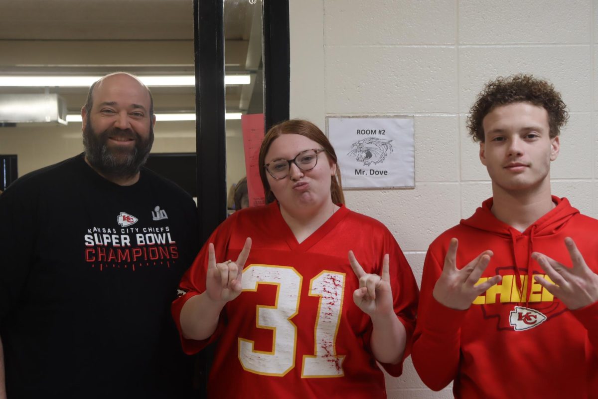 History teacher Matt Dove and Sophomores Mya Taylor and DAngelo Ramirez stand outside Doves classroom posing with their Chiefs gear. They are hopeful for a Chiefs on Sunday. 
